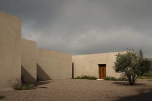 Modern rammed earth house exterior with wooden door, olive tree, and gravel courtyard under a stormy sky