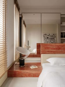 Modern bedroom featuring a raised red marble platform bed with white bedding, a sculptural white and black lamp, wooden blinds, and black-and-white artwork on the wall.
