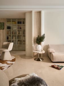 Modern living room featuring a travertine-topped pedestal side table with a potted plant, a beige swivel armchair on a cowhide rug, built-in floor-to-ceiling bookshelves, and a curved beige sofa under warm natural light.