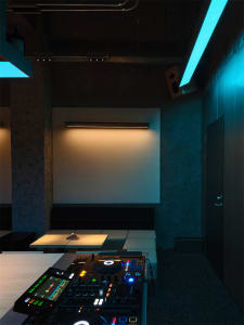 Interior view of a modern DJ booth featuring a Pioneer DJ mixer and turntables in the foreground, set against raw concrete walls with linear LED lighting in cyan and warm white tones.