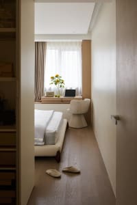 View into a minimalist bedroom featuring a low-profile bed with white linens, a boucle swivel chair at a built-in window desk, sheer curtains, and light wood flooring.