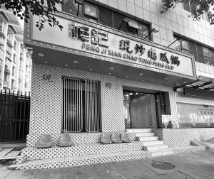 Black and white photograph of a retro-style restaurant storefront with black and white mosaic tile walls, four plastic stools on the steps, vertical metal security gate, and large illuminated sign reading Feng Ji Xian Chao Tong Feng Guo.