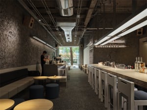 Long narrow industrial-style cafe interior with textured concrete walls, a wooden bar counter with white stools on the right, built-in booth seating on the left, round wooden tables, and an open view to greenery at the far end.