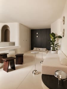Modern minimalist living room featuring a cream boucle sofa, built-in beige daybed with pillows, dark wood stools, marble-topped side tables, and a large potted plant against a dark wood accent wall.