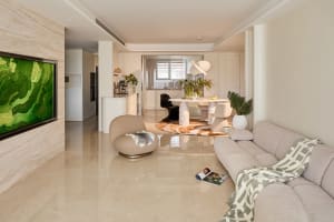 Modern open-plan living room and dining area with travertine marble feature wall, large green abstract artwork, light beige sectional sofa, sculptural white marble dining table, and glossy reflective floor.