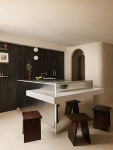 Modern minimalist kitchen featuring a walnut and stainless steel island with white tiled overhang and dark wooden block stools in a bright, neutral space with an arched doorway.