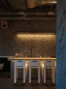 Modern industrial bar counter with three white stools, concrete feature wall, and under-cabinet lighting in a loft-style space.