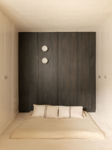 Minimalist Japanese-style bedroom featuring a floor-level tatami bed with beige linen bedding, a large dark wood paneled wall, and two spherical white wall sconces.