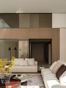 Modern minimalist living room featuring light oak wood paneling, two neutral fabric sofas, round coffee tables, and a glass vase with yellow forsythia branches.