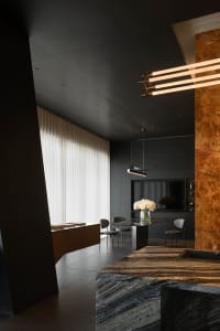 Modern dark interior featuring a large marble kitchen island in the foreground, a dining table with white flower arrangement, dramatic linear pendant lights, and a tall window with sheer curtains.