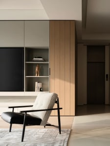 Modern living room featuring a black and white boucle armchair with black metal frame on a textured rug, positioned in front of built-in light wood and off-white cabinetry with open shelving and a large television.