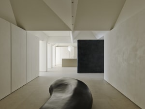 Long minimalist hallway with off-white micro-cement walls and floor, a large amorphous black sculptural bench in the foreground, and a dramatic dark textured accent wall at the end.