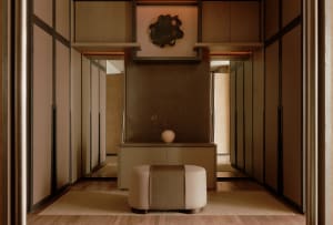 Modern minimalist entryway featuring a walnut console cabinet, upholstered ottoman with woven center band, dark metal leaf wall art, and warm ambient lighting in a neutral palette.