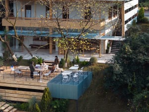 Aerial view of a modern mountain villa featuring a large wooden deck with dining area, a striking blue-green corten steel platform, and people enjoying the outdoor space surrounded by trees.