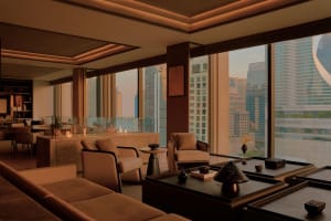 Modern luxury living room featuring a linear travertine fireplace, beige upholstered sofas, dark wood coffee tables, and floor-to-ceiling windows showcasing a dense city skyline at dusk.