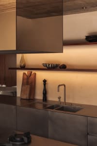 Modern minimalist kitchen featuring a brushed stainless steel countertop and double sink, dark cabinetry, floating walnut open shelf with decorative objects, and warm ambient lighting.