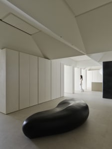 Minimalist gallery interior featuring a large black organic-shaped sculpture in the foreground, floor-to-ceiling micro-cement walls, and a woman in white standing in the distance under dramatic angular ceilings.