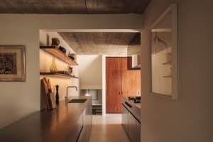 Modern minimalist kitchen featuring warm walnut wood cabinetry, concrete ceiling, stainless steel countertops, and open shelving with curated ceramics.