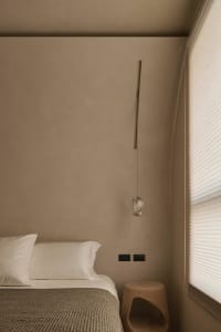 Minimalist bedroom corner featuring a textured travertine wall, white bedding, a hanging crystal pendant light, and a solid wood stump stool beside a window with sheer blinds.