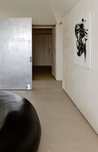 Modern minimalist hallway featuring seamless micro-cement walls, concrete floor, a large black sculptural object in the foreground, and a black and white abstract engine artwork hanging on the right wall.