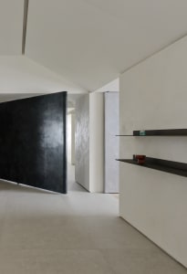 Modern minimalist hallway featuring a large black pivoting door, light micro-cement walls, floating black shelves with decorative objects, and seamless concrete flooring.