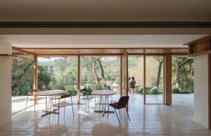 Bright modern indoor-outdoor dining area with round wooden tables, mixed-style chairs, large wood-framed glass doors opening to a lush green garden and pond.