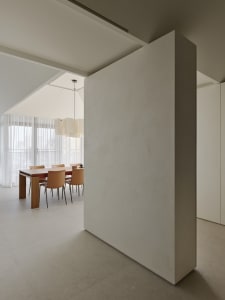 Modern minimalist dining room featuring a large micro-cement partition wall, wooden dining table, tan leather chairs, and a sculptural pendant light under a high sloped ceiling.