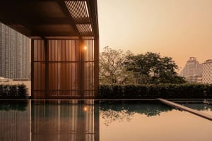 Modern rooftop infinity pool reflecting a dramatic orange sunset, framed by vertical wooden slat screens and a city skyline in the background.