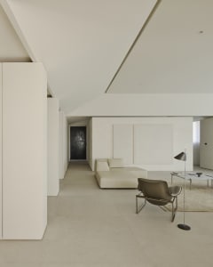 Minimalist living room featuring off-white micro-cement floors and walls, a low-profile cream leather sectional sofa, a dark olive sculptural lounge chair with chrome frame, and a slim floor lamp.