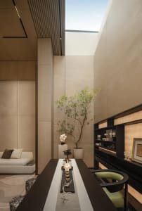 Modern minimalist dining room featuring travertine walls, a long dark wood table, built-in dark shelving, and a tall potted tree under a large skylight.