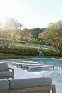 Modern infinity pool with teak and fabric lounge chairs on a stone deck, a person tending to plants in the background with forested hills beyond.
