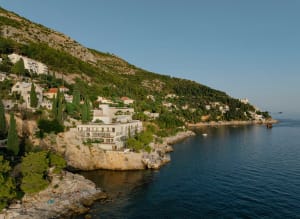 A modern white cliffside hotel built into rocky terraces overlooking the deep blue Adriatic Sea, with people sunbathing on the rocks below and green hills rising behind.