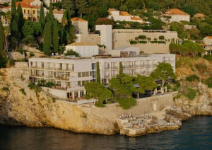 Modern white luxury hotel built into a rocky cliffside with multiple terraces, stone retaining walls, and direct access to the sea below.
