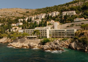 A contemporary seaside hotel built directly on a rocky coastline with turquoise water in the foreground and a hillside village rising behind it under a clear blue sky.