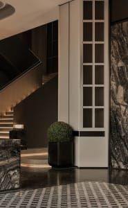 Modern luxury entrance hall featuring black marble floors and walls, a white paneled wall with grid windows, a spherical topiary in a black planter, and a floating staircase with warm LED step lighting.
