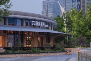 Modern curved restaurant exterior with stone base, large glass windows, and illuminated signage at dusk, surrounded by trees and city buildings in the background.