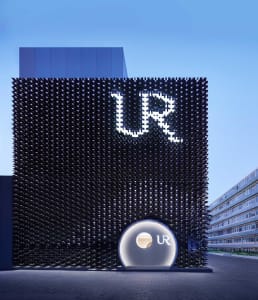 Night view of a modern building with a black perforated metal facade featuring a large illuminated UR logo and a circular entrance structure.