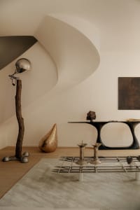 Modern minimalist living room featuring a sculptural black console table, a floor lamp made from a real tree trunk with a vintage helmet shade, a textured gold teardrop sculpture, and a glass coffee table with metallic candle holders on a neutral rug.