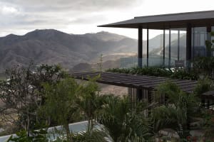 Modern mountain villa with large glass windows and a dark wooden terrace overlooking a vast tropical mountain valley under cloudy skies.