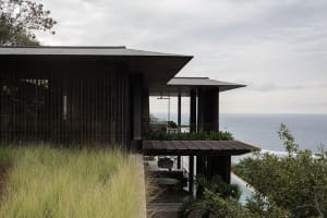 Modern dark wood villa with large cantilevered terrace overlooking the ocean, surrounded by tall green grasses under an overcast sky.