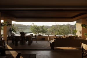Modern open-plan living and dining area with large sofa, round dining table, and expansive view of a golf course and tropical landscape.