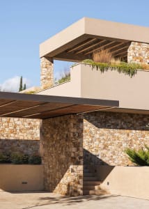 Modern Mediterranean outdoor terrace featuring stacked natural stone walls, a floating concrete pergola with wooden slats, and lush rooftop planting under a clear blue sky.