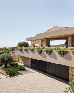 Modern Mediterranean villa exterior with stone walls, concrete overhang, cascading rooftop garden, and paved courtyard under bright sunlight.