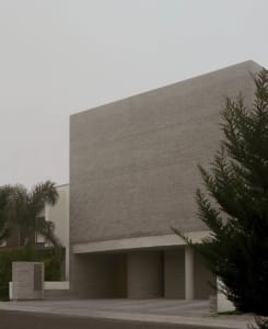 Exterior view of a brutalist concrete brick building with a large overhanging rectangular volume and wooden entrance door.