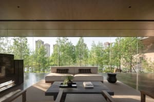 Modern minimalist living room featuring a low-profile cream sofa, dark stone coffee table, wood-paneled ceiling, and expansive floor-to-ceiling windows overlooking lush green trees and city buildings.