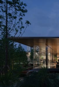 Modern house with floor-to-ceiling glass walls opening to a tropical garden and wooden deck at twilight, showing warm interior lighting and seamless indoor-outdoor connection.