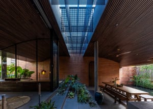 Modern tropical indoor courtyard with exposed red brick walls, dark wooden slat ceiling, central greenery, long wooden dining table, and a large skylight with perforated panels.