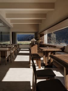 Modern rustic dining room interior with solid wood tables and chairs, concrete floor, large windows overlooking mountains and grassland.