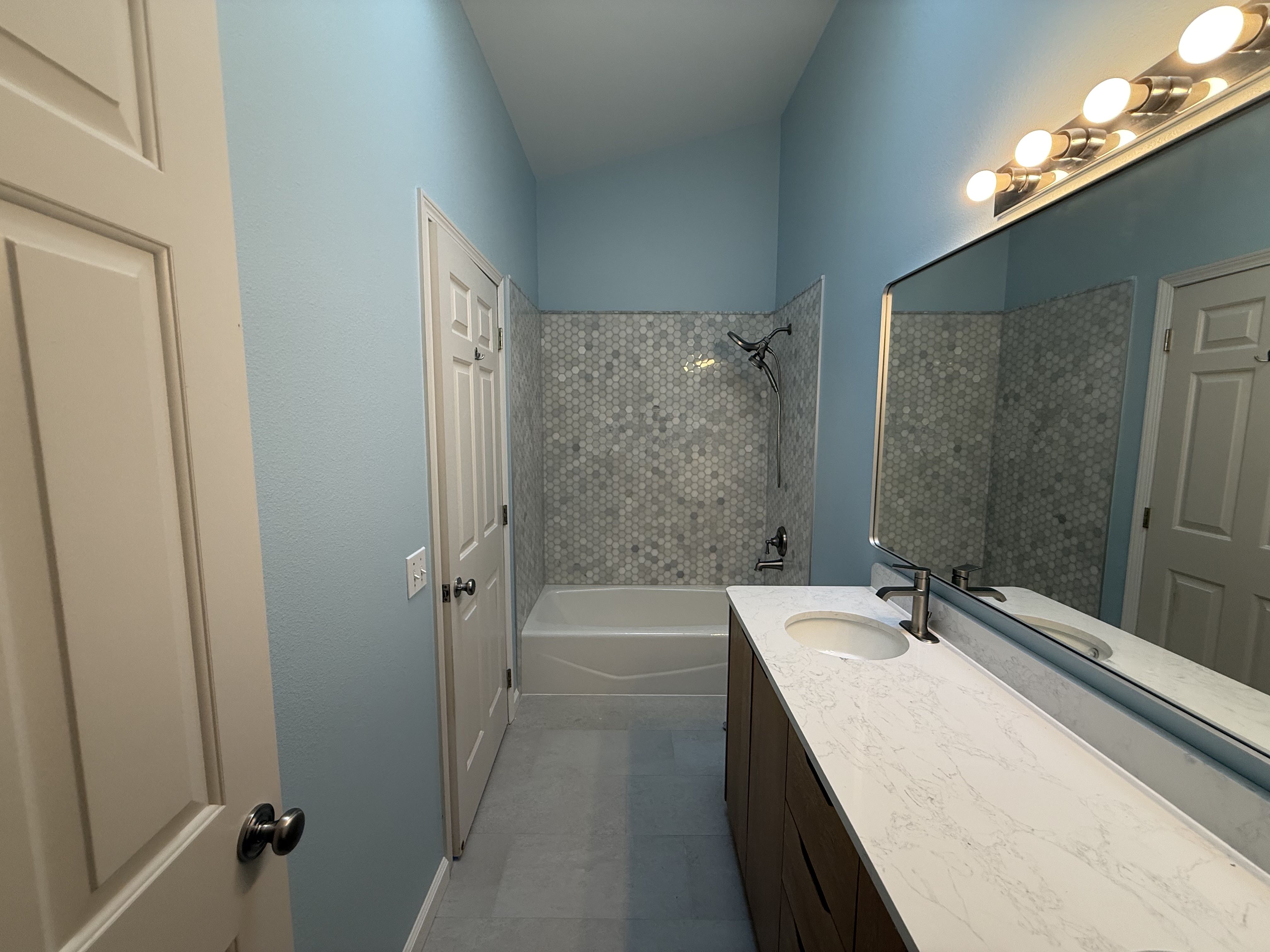 After: oak slab-front vanity, white quartz top and backsplash, dual brushed-nickel faucets, large rounded-corner mirror; hex mosaic at tub; light-gray porcelain floor