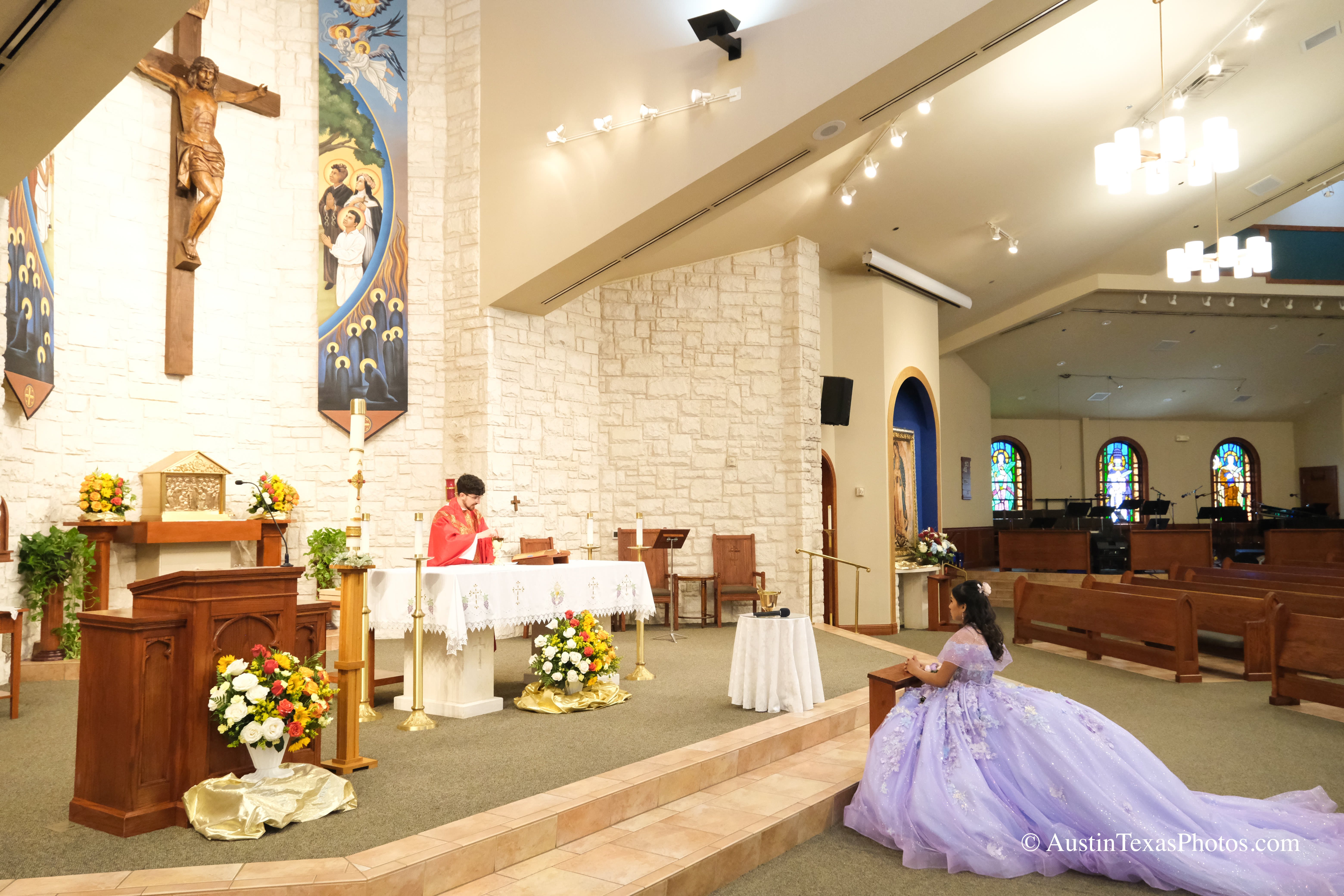 Quinceañera in lavender gown kneeling at a prayer bench during mass at Santa Cruz Catholic Church, Buda, Texas, with limestone sanctuary walls and altar behind her